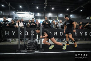 Yésica Herrera ejecuta el exigente empuje de trineo (Sled Push) bajo la atenta mirada y el aliento de su compañero de equipo, José Pablo Leoni, durante la prueba en Buenos Aires. Foto: Hyrox Buenos Aires.